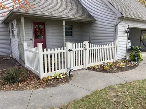 White picket fence and gate enclosing a front porch beside a garden bed.