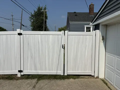 Pale woodgrain-look privacy gate with dark hinges and latch beside a garage.